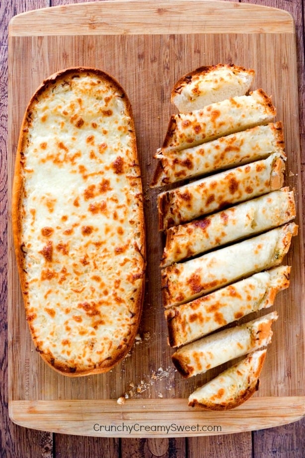 Overhead shot of cheesy garlic bread on wooden cutting board, one half sliced and the second full.