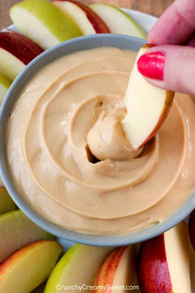 Caramel cheesecake dip with hand dipping apple slice in it.