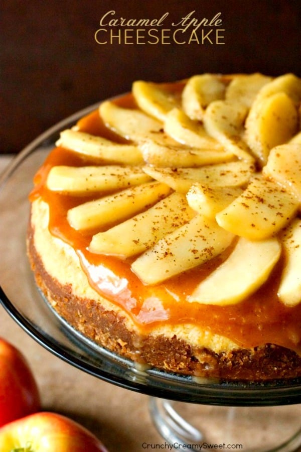 Side shot of cheesecake with apple slices and caramel sauce on top, on glass cake stand.