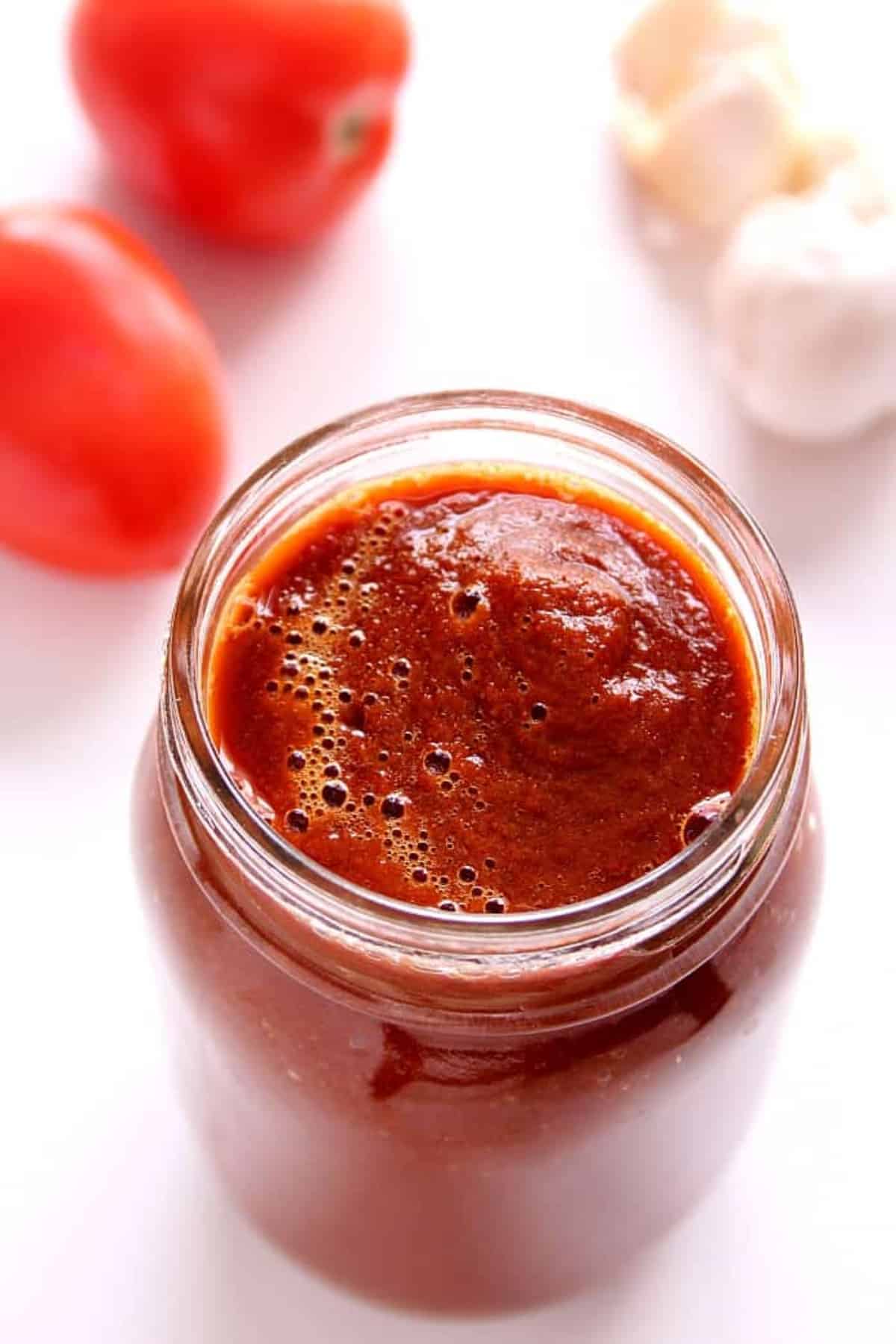 Red enchilada sauce in a glass jar on a white board with tomatoes and garlic next to it.