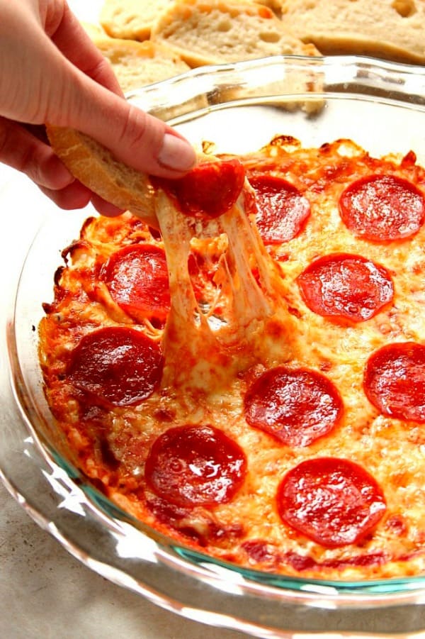 Pepperoni Pizza Dip in glass pie dish, baguette slice being dipped into cheese.