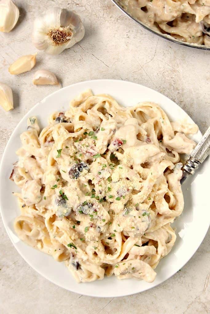 Overhead shot of mushroom chicken Alfredo pasta on white plate, garlic cloves next to it.