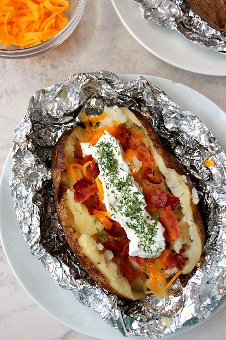 Overhead shot of baked potato in foil, topped with bacon, cheese and sour cream, set on white plate.