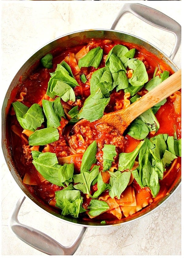 Overhead shot of tomato sauce with sausage, lasagna noodles and fresh spinach on top in skillet.