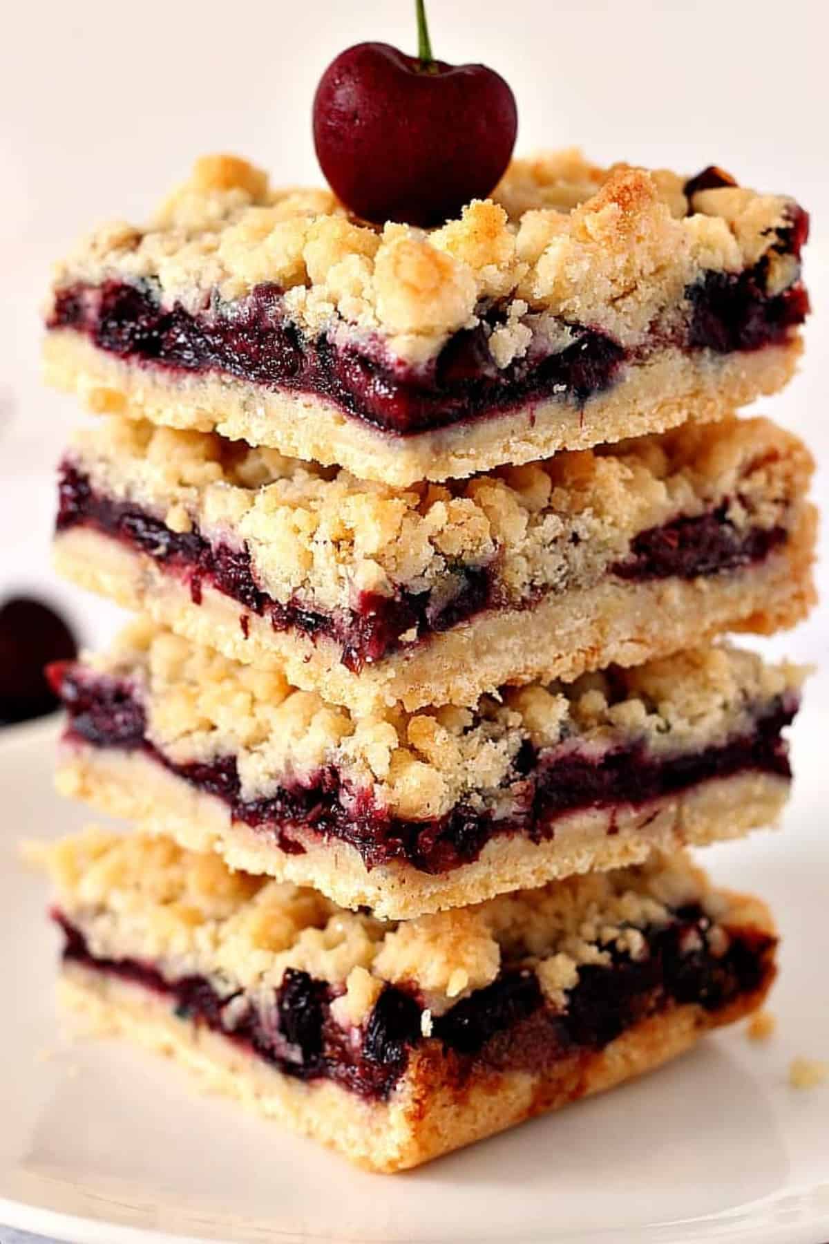 Four square pieces of cherry pie crumb bars stacked on a white plate with a fresh cherry on the top square.