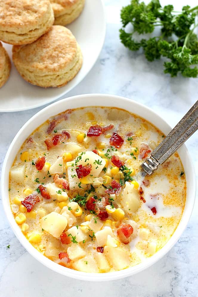 Overhead shot of corn chowder with potatoes and bacon in a white bowl, with plate of biscuits next to it.