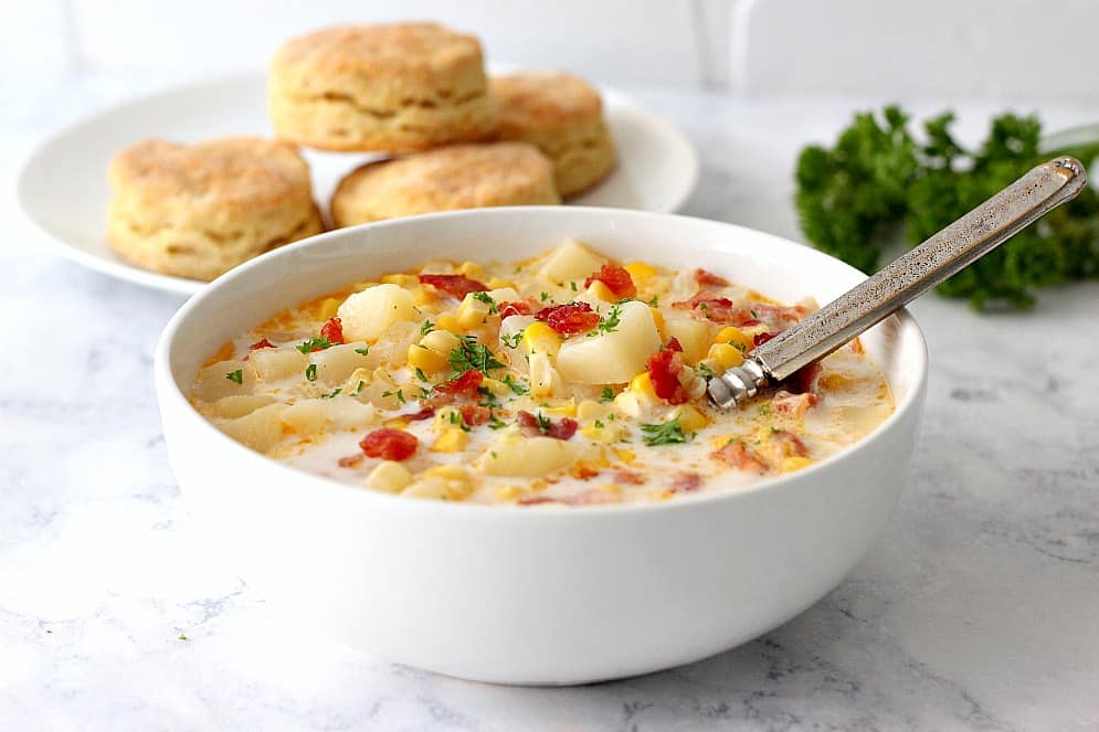 Side shot of white bowl with corn chowder with biscuits in the background.