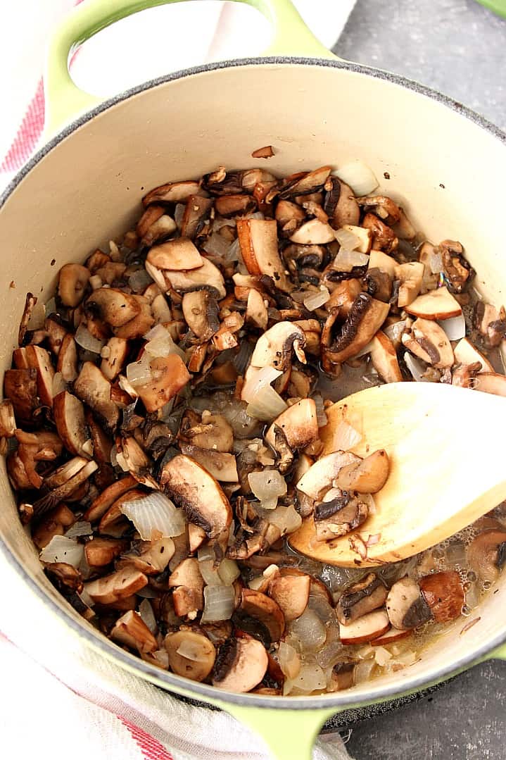 Sauteed mushrooms and onions in a green Dutch oven with a wooden spoon.