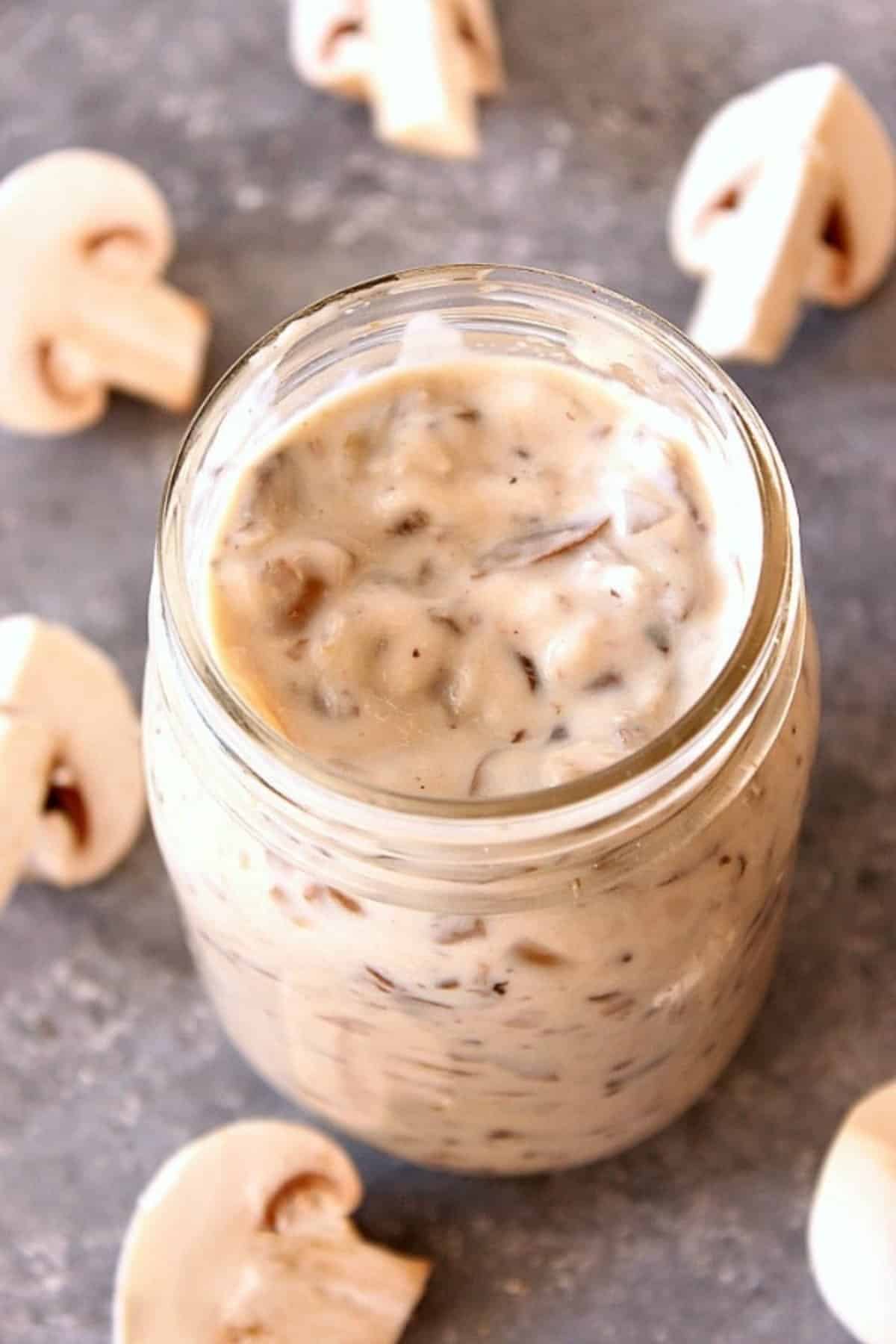 Cream of mushroom soup in a glass Mason jar, on a gray board with mushrooms around it.