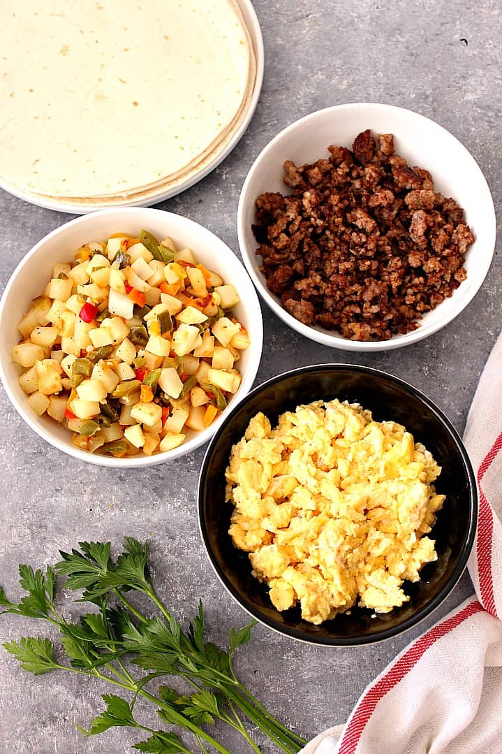 Overhead shot of potatoes in bowl, scrambled eggs in bowl and cooked sausage in bowl, next to tortillas.