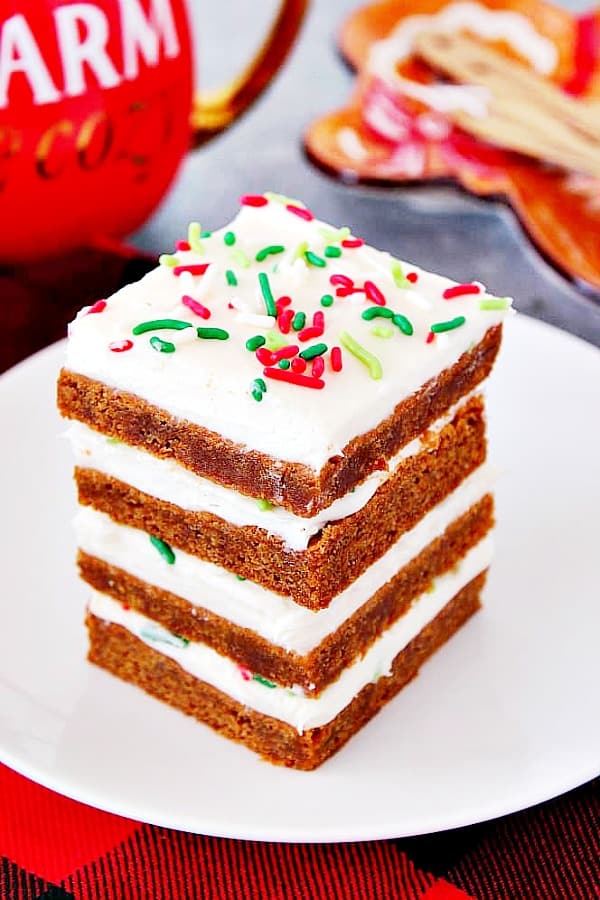 These Gingerbread Bars are chewy, perfectly spiced cookie bars topped vanilla cream cheese frosting and festive sprinkles. Gingerbread Bars with frosting stacked on white plate.