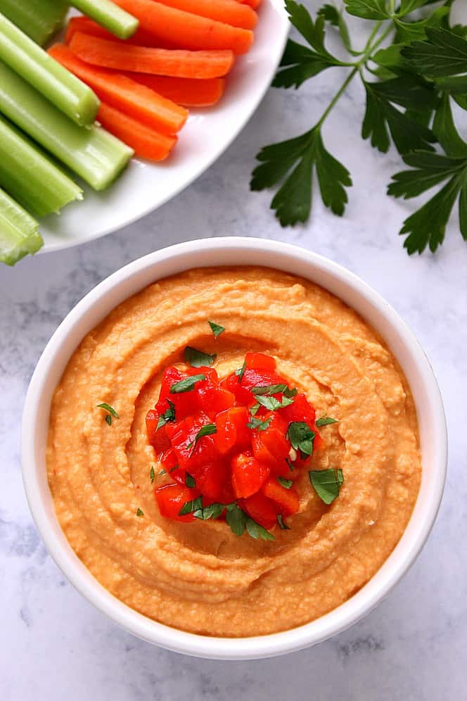 Roasted Red Pepper Hummus in a white bowl on marble board with parsley and veggie sticks on the side.