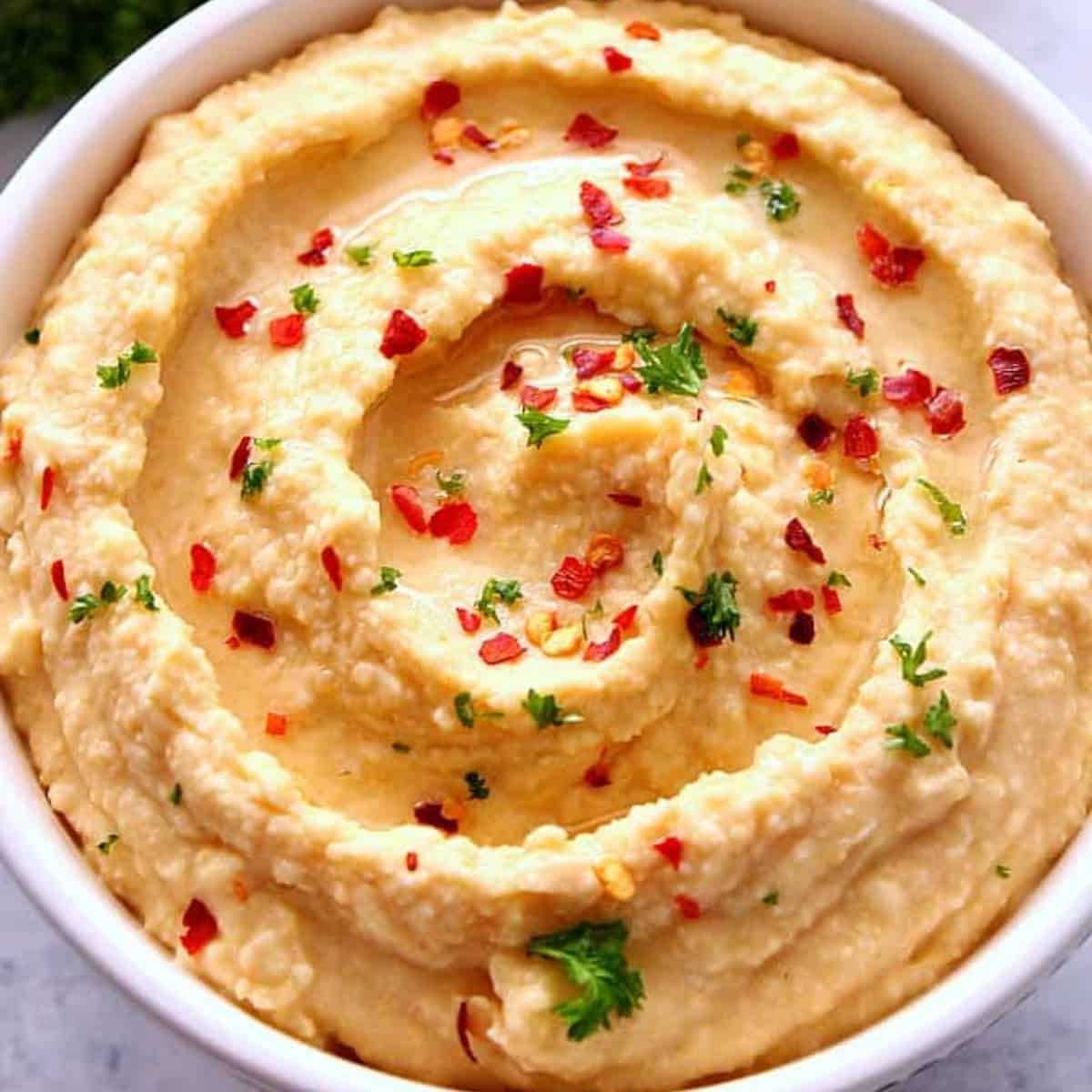 Square image of hummus with red pepper flakes in a bowl, on a marble board.