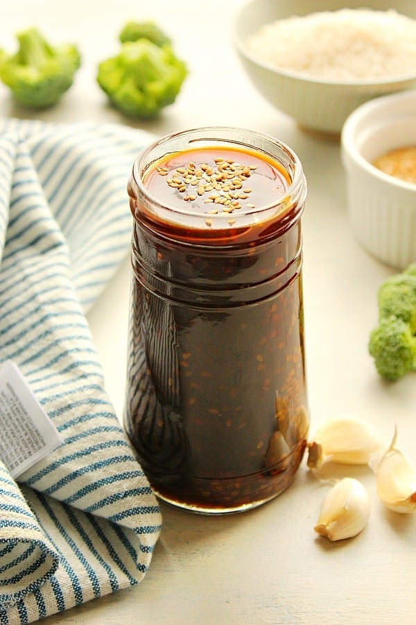 Teriyaki Sauce in a jar with sesame seeds on top, set on a white board with kitchen towel next to it.