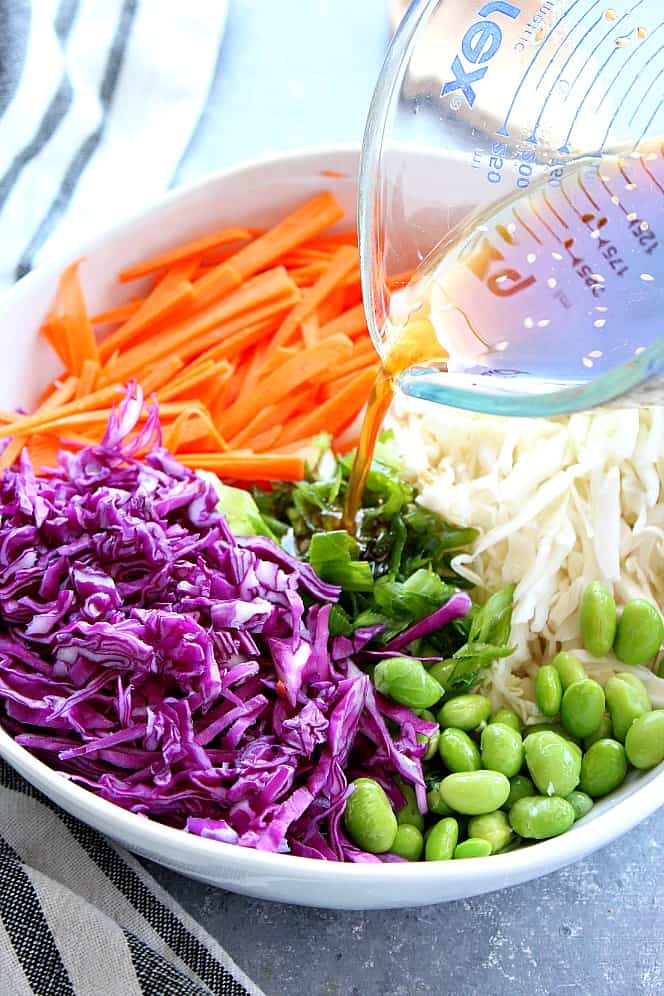 Close up on bowl with purple cabbage, carrots, green cabbage, edamame beans and dressing pouring from a glass cup. 