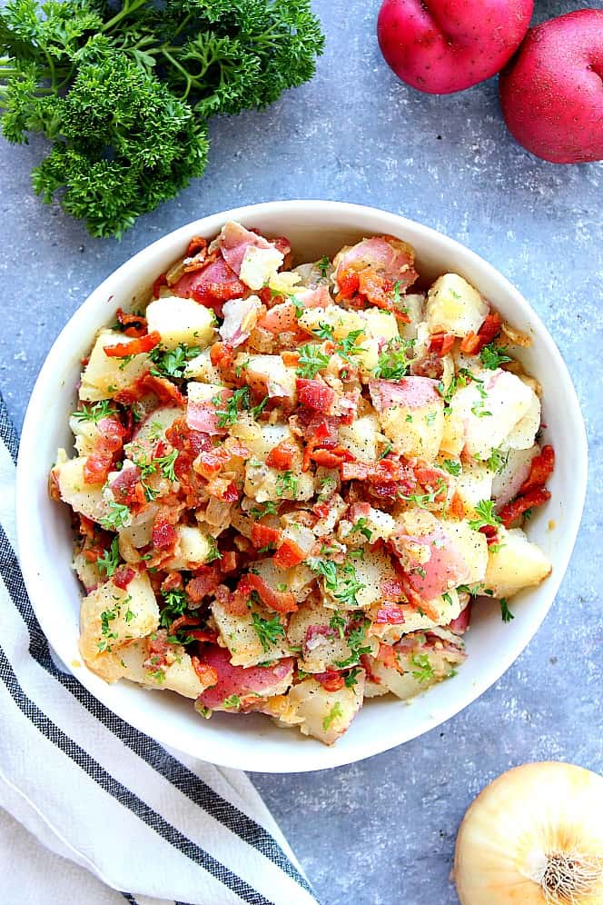 Overhead shot of potato salad served in a white bowl, wish parsley and red potatoes in the background.