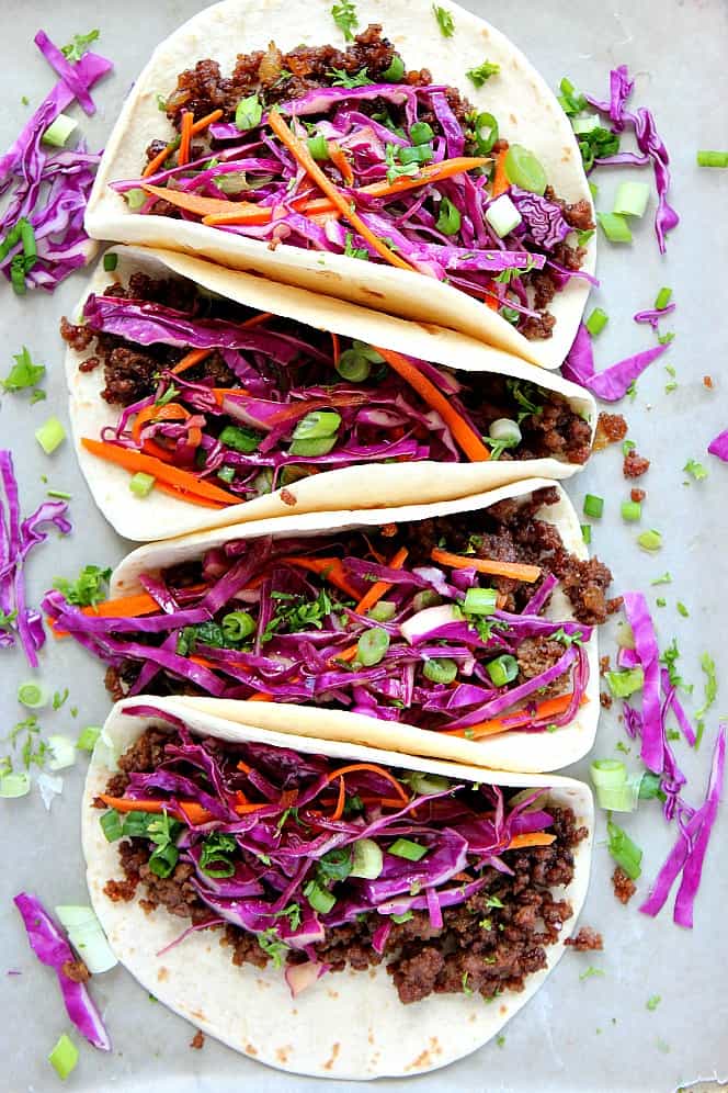 Four tacos made with soft tortillas, filled with Korean beef and topped with Asian slaw, presented on baking sheet. 