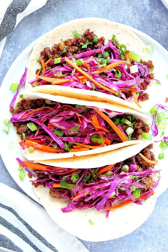 Korean Beef Tacos - ground beef caramelized in sweet soy sauce paired with Asian Slaw in flour tortillas. #Asian #beef Three Korean Beef Tacos with Asian slaw on white plate.