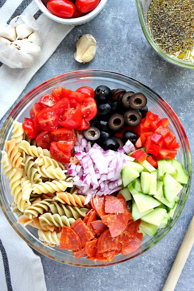 Overhead shot of pasta, tomatoes, olives, cucumber, pepperoni, red pepper and onion in a mixing bowl. 