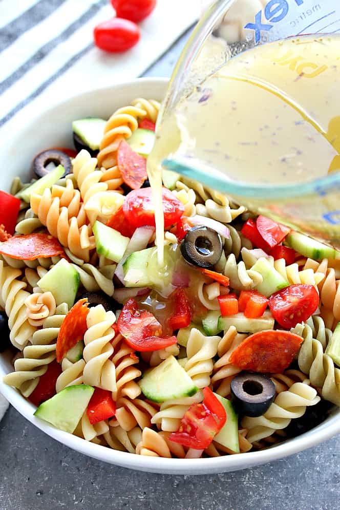 Italian pasta salad in a bowl, with dressing being poured from a measuring cup. 