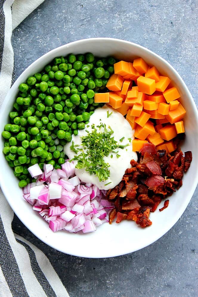 Green peas, red onion, cheddar, bacon and dressing in a white bowl, before tossing for a salad.