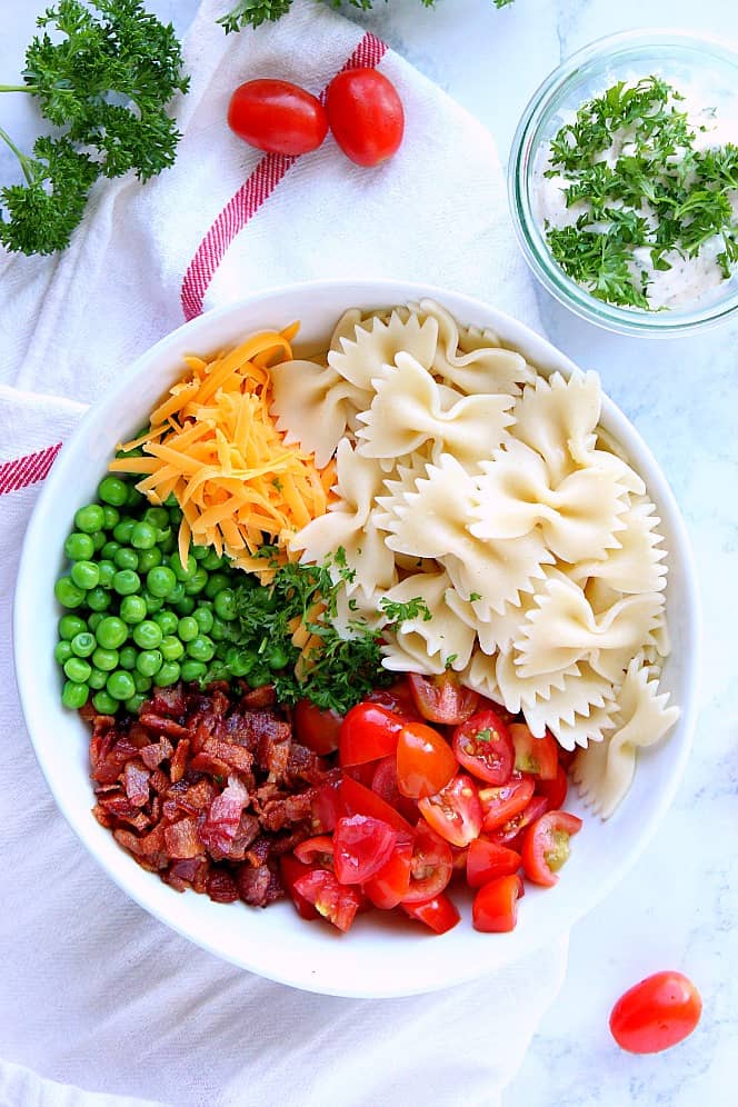 Overhead shot of salad ingredients: pasta, cheddar, peas, bacon and cherry tomatoes in a white bowl on white towel.