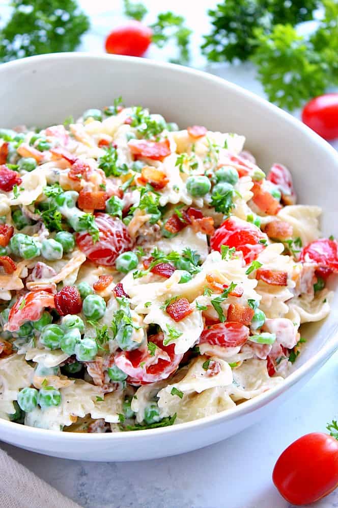 Close up photo of pasta salad with bacon, peas and tomatoes in a white bowl. 