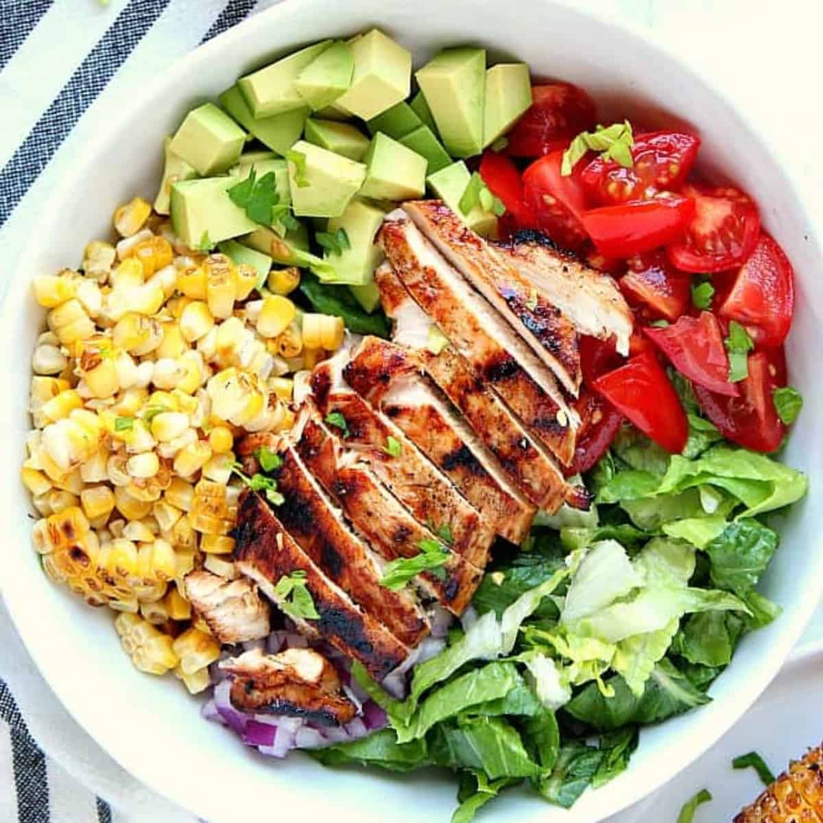 Square image of salad with grilled chicken, corn, tomatoes and avocado in a white bowl.