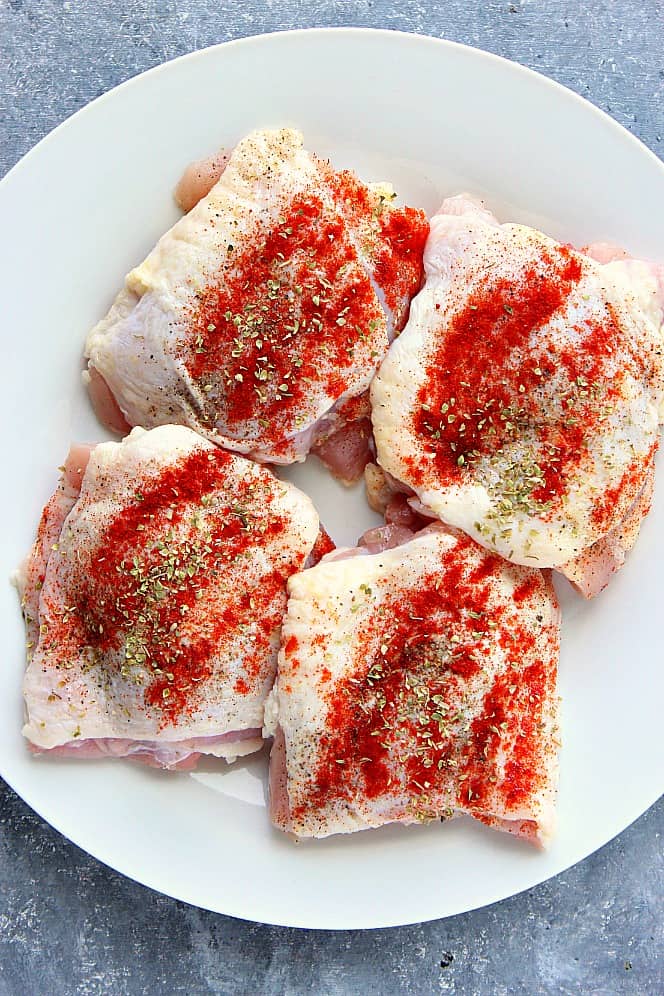 Overhead shot of raw bone-in skin-on chicken thighs, seasoned with salt, pepper, paprika and oregano, on white plate. 