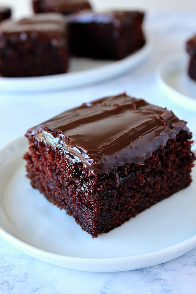 Side shot of a piece of chocolate cake with fudgy chocolate frosting, on white plate with other plates in background.