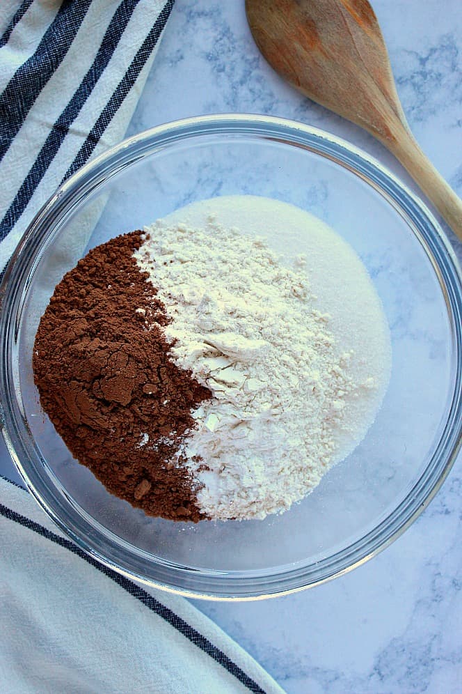 Overhead shot of flour, cocoa powder and sugar in a mixing bowl, with wooden spoon next to it.