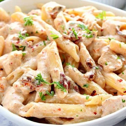 Square image of penne pasta with chicken in sun-dried tomato sauce in a white pasta bowl.