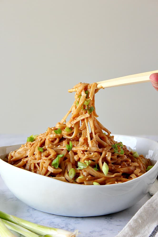 Side shot of white bowl filled with peanut noodles lifted by chopsticks. 