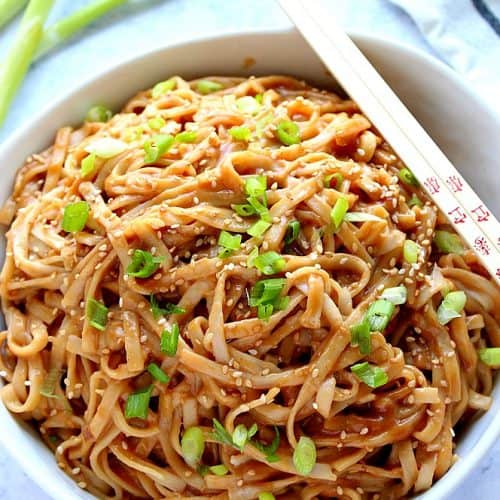 Noodles with peanut sauce and green onions in a white bowl with chopsticks.