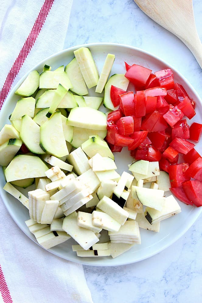 Overhead shot of chopped zucchini, eggplant and tomatoes on white plate. 