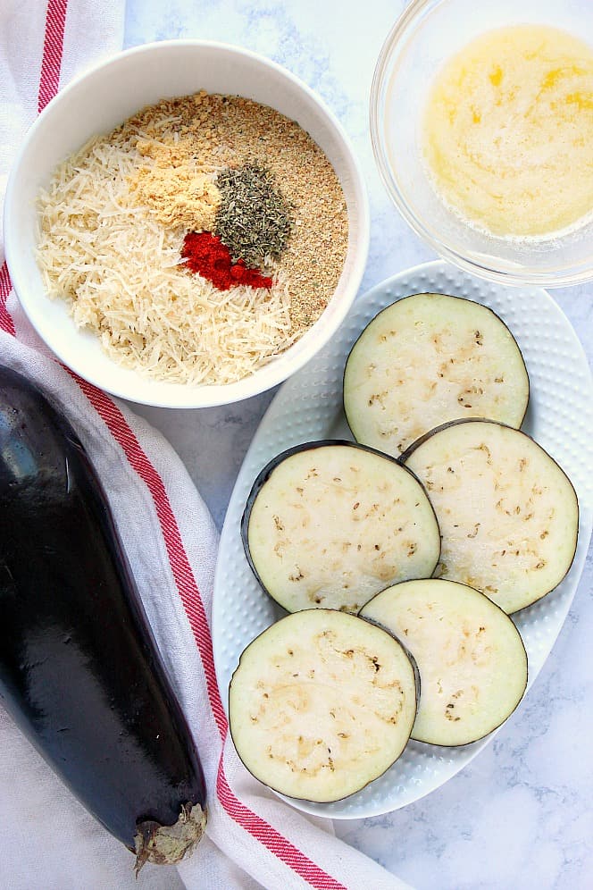 Overhead shot of sliced eggplant in oval bowl, breadcrumbs with spices in white bowl and melted butter in glass bowl. 