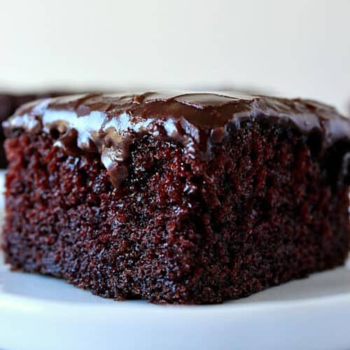 Square image of a piece of chocolate cake with shiny fudge frosting, set on a white dessert plate.