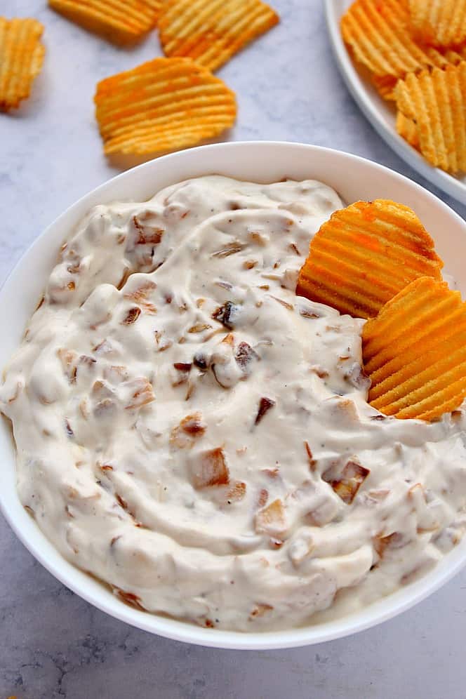 Close up shot of caramelized onion dip in white bowl with cheddar ripple potato chips on side. 