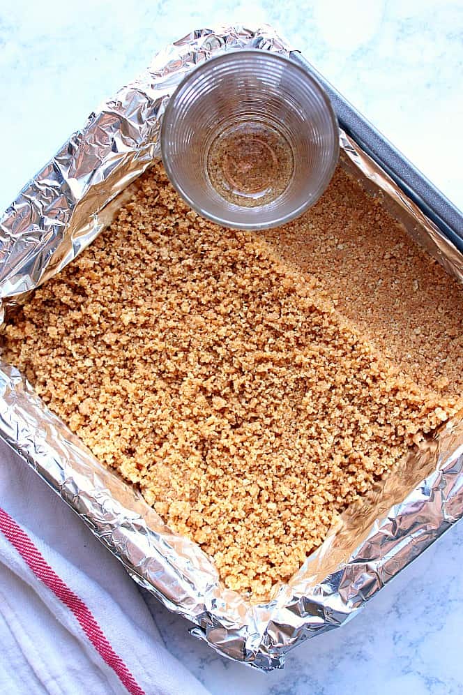 Overhead shot of graham cracker crust being pressed onto the bottom of baking pan with glass. 