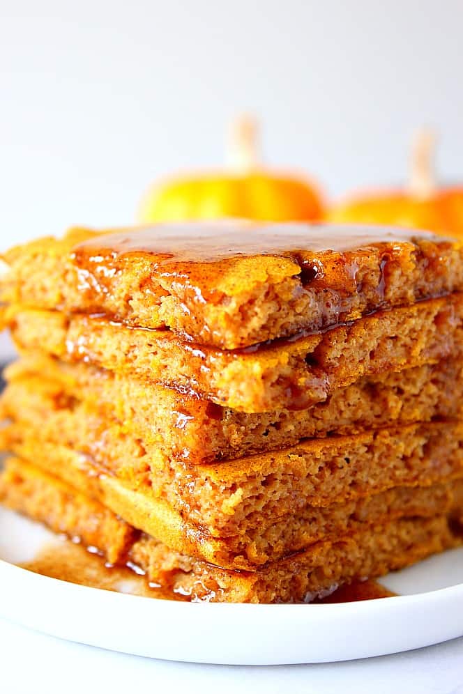 Pumpkin Sheet Pan Pancakes Recipe - soft, fluffy and filled with pumpkin and pumpkin spice pancakes baked in the oven on a sheet pan. No more flipping! Serve with spiced brown sugar butter. Close up shot of pumpkin pancakes stacked on white plate, with a drizzle of melted butter.