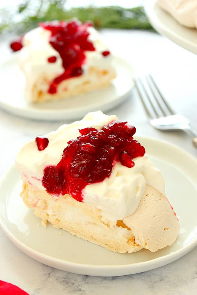 Side shot of a slice of Pavlova cake on white plate, topped with whipped cream and cranberry sauce. 