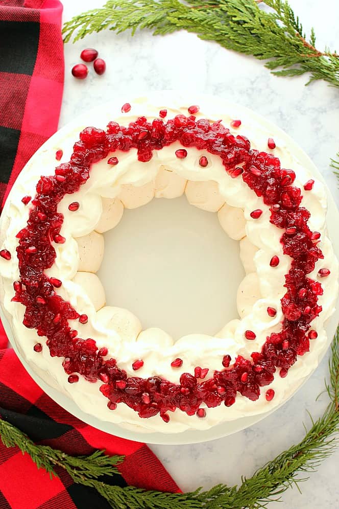 Overhead shot of Pavlova cake shaped like a wreath, topped with whipped cream, cranberry sauce and pomegranate. 