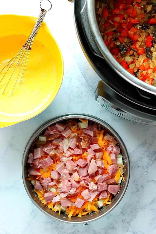 Overhead shot of Instant Pot with sauteed veggies, egg mixture in yellow mixing bowl and cheese and ham layer in springform pan. 