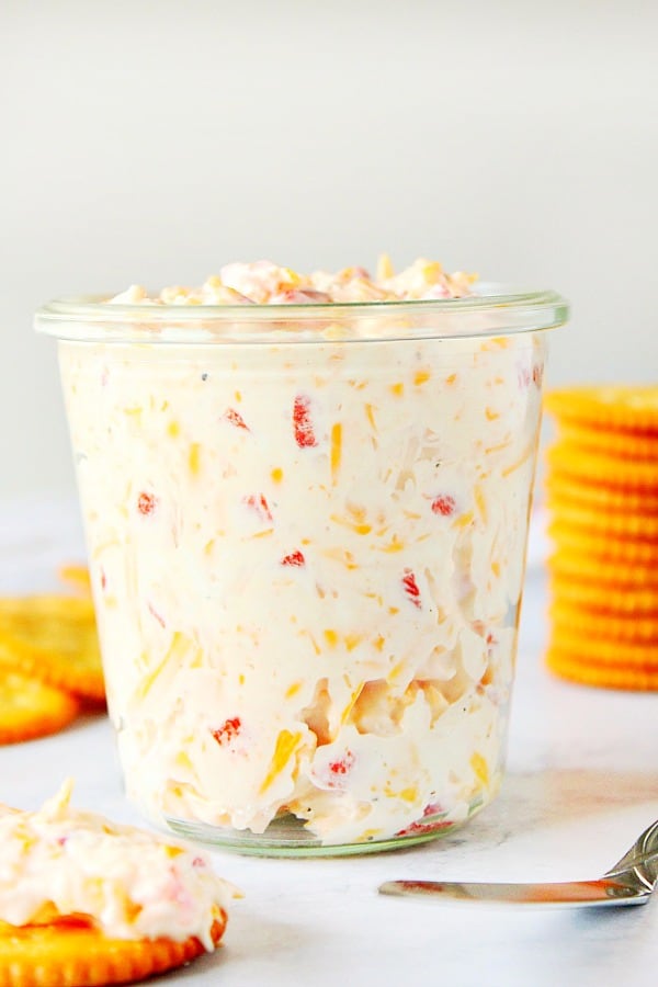 Pimento Cheese Spread Recipe made with chopped pimento peppers, shredded cheddar cheese, cream cheese and mayo. Quick and easy as cracker topping or sandwich filling for your next party! Pimento Cheese Spread in a glass jar, with crackers behind it.