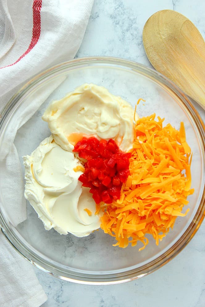 Overhead shot of cheddar, mayo, cream cheese and chopped pimento peppers in glass mixing bowl.