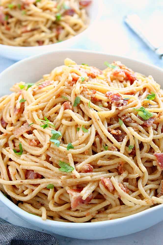 Side shot of spaghetti carbonara with bacon and chopped parsley in large white serving bowl. 