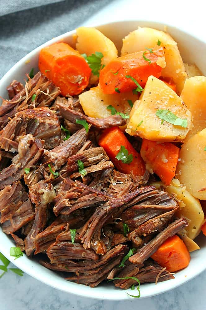 Overhead shot of shredded pot roast with carrots and potatoes on the side in white bowl. 