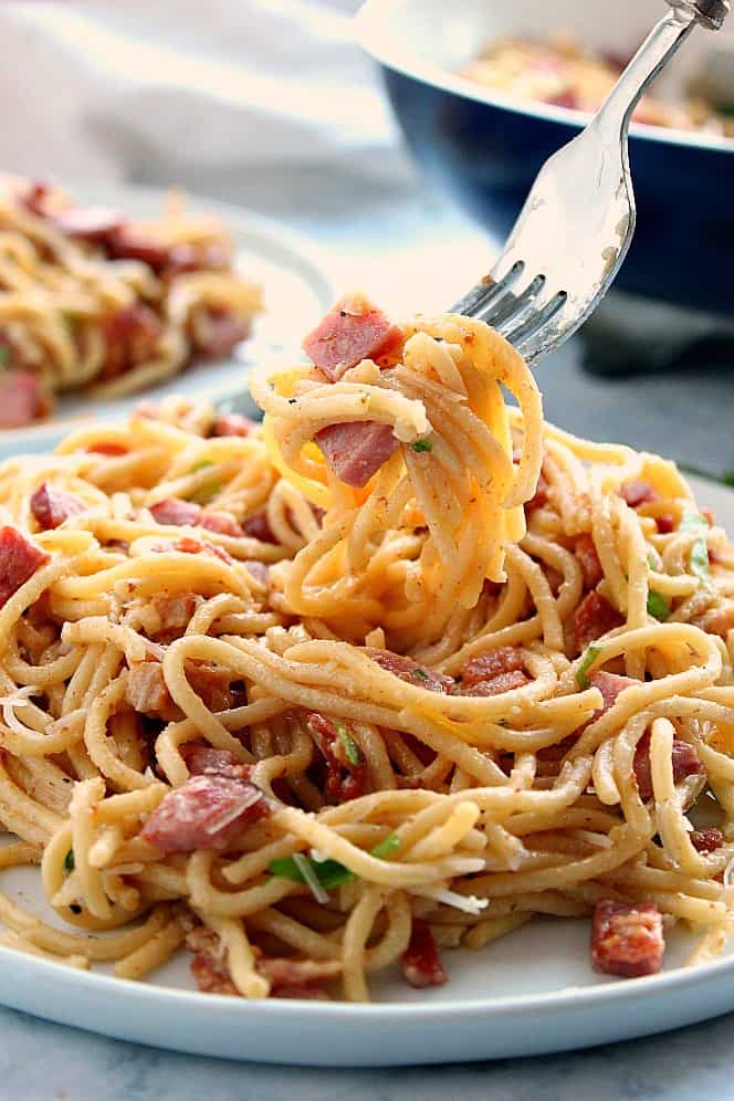 Close side shot of spaghetti carbonara with ham on white plate, some noodles on a fork.