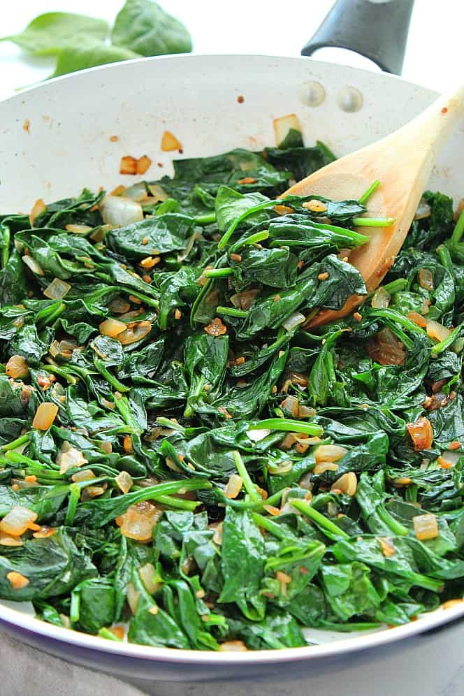 Side clos up shot of sauteed spinach with onion in large white pan.