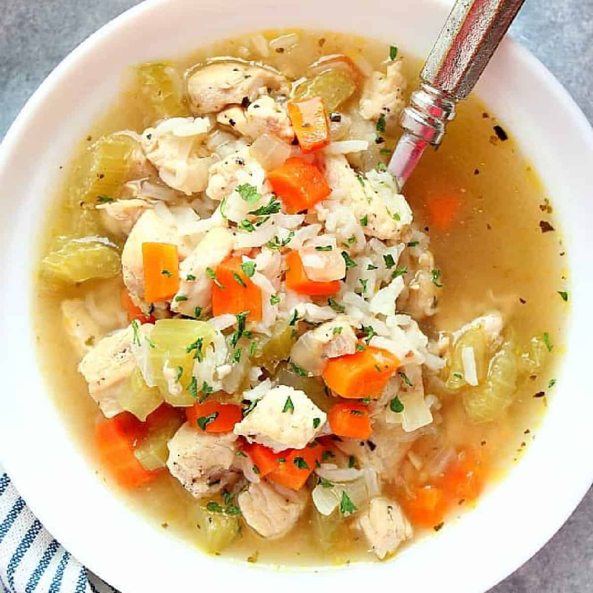 Square image of chicken and rice soup in a white bowl with silver spoon.